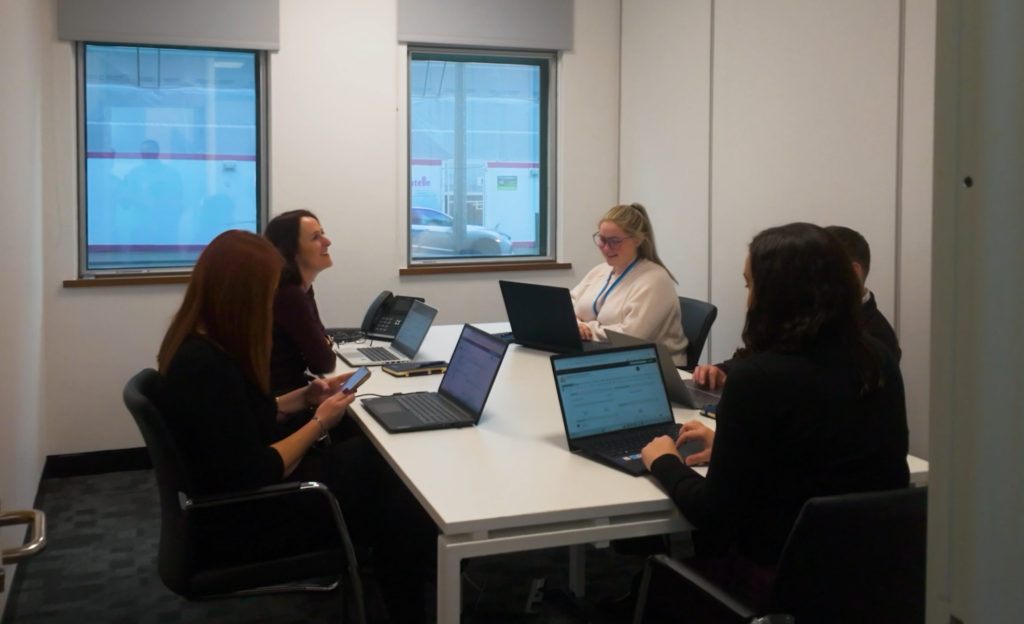 The Howells Team sits happily around a table in a meeting room.